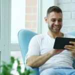 Happy man sitting in blue chair indoors, using a tablet, bright natural lighting.