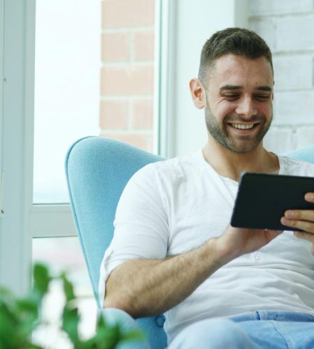 Happy man sitting in blue chair indoors, using a tablet, bright natural lighting.